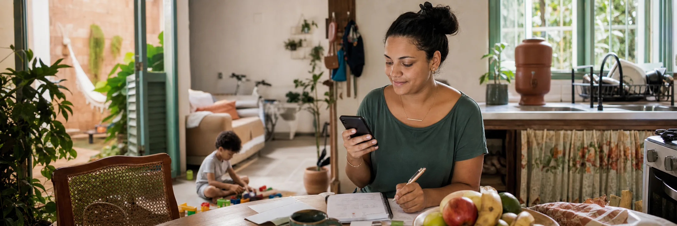 Mãe brasileira usando o celular durante a rotina da casa, com uma criança brincando por perto.